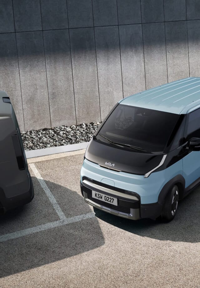 Der Kia PV5 Passenger fährt aus einer Parallelparklücke – auf einem Parkplatz mit zwei weiteren Kleinbussen.